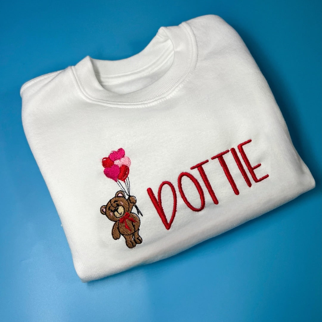 White sweatshirt with teddy bear and balloons design and 'DOTTIE' text on a blue background