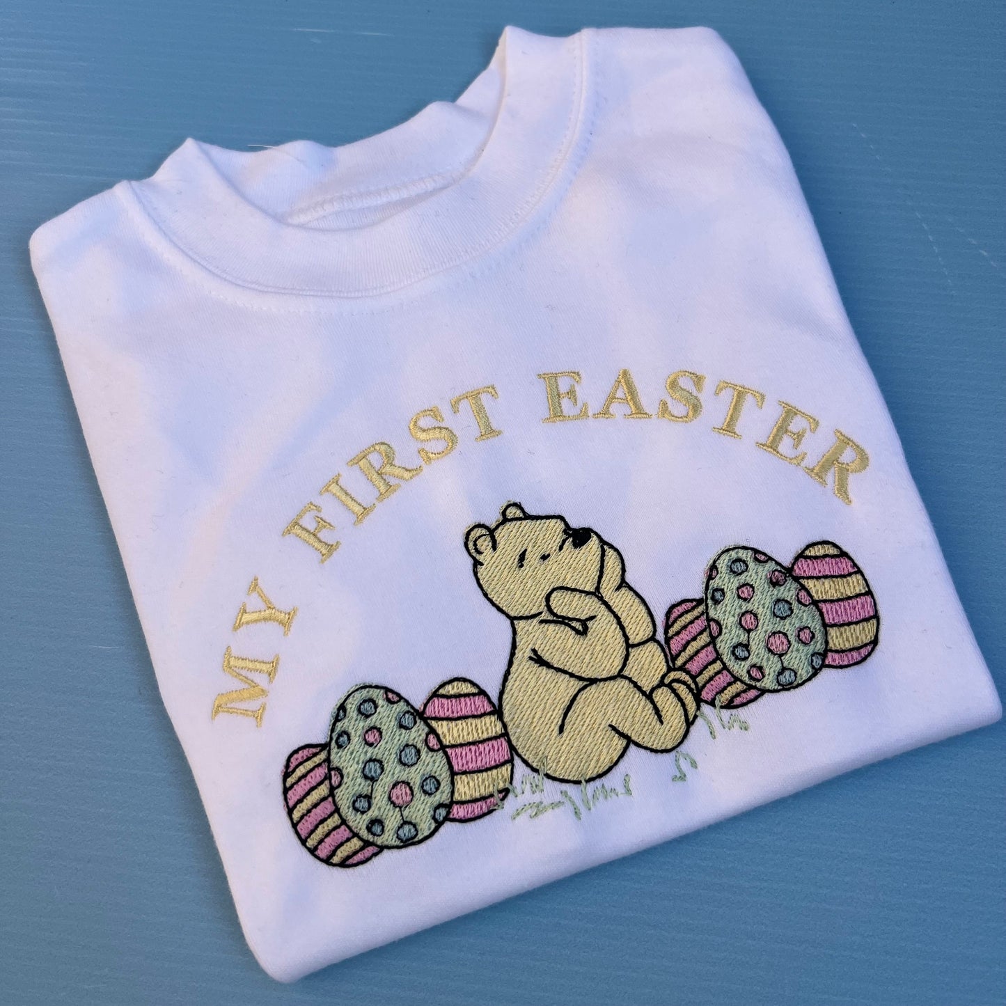 Folded white shirt with 'My First Easter' text and bear holding eggs on a blue background