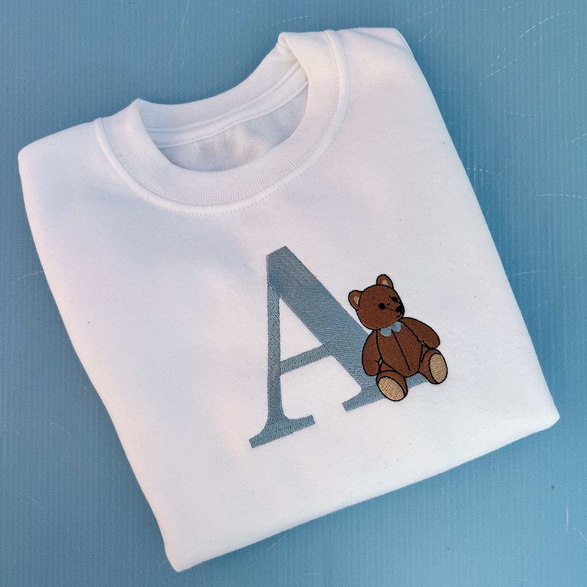 White t-shirt with a brown teddy bear and letter 'A' on a blue background