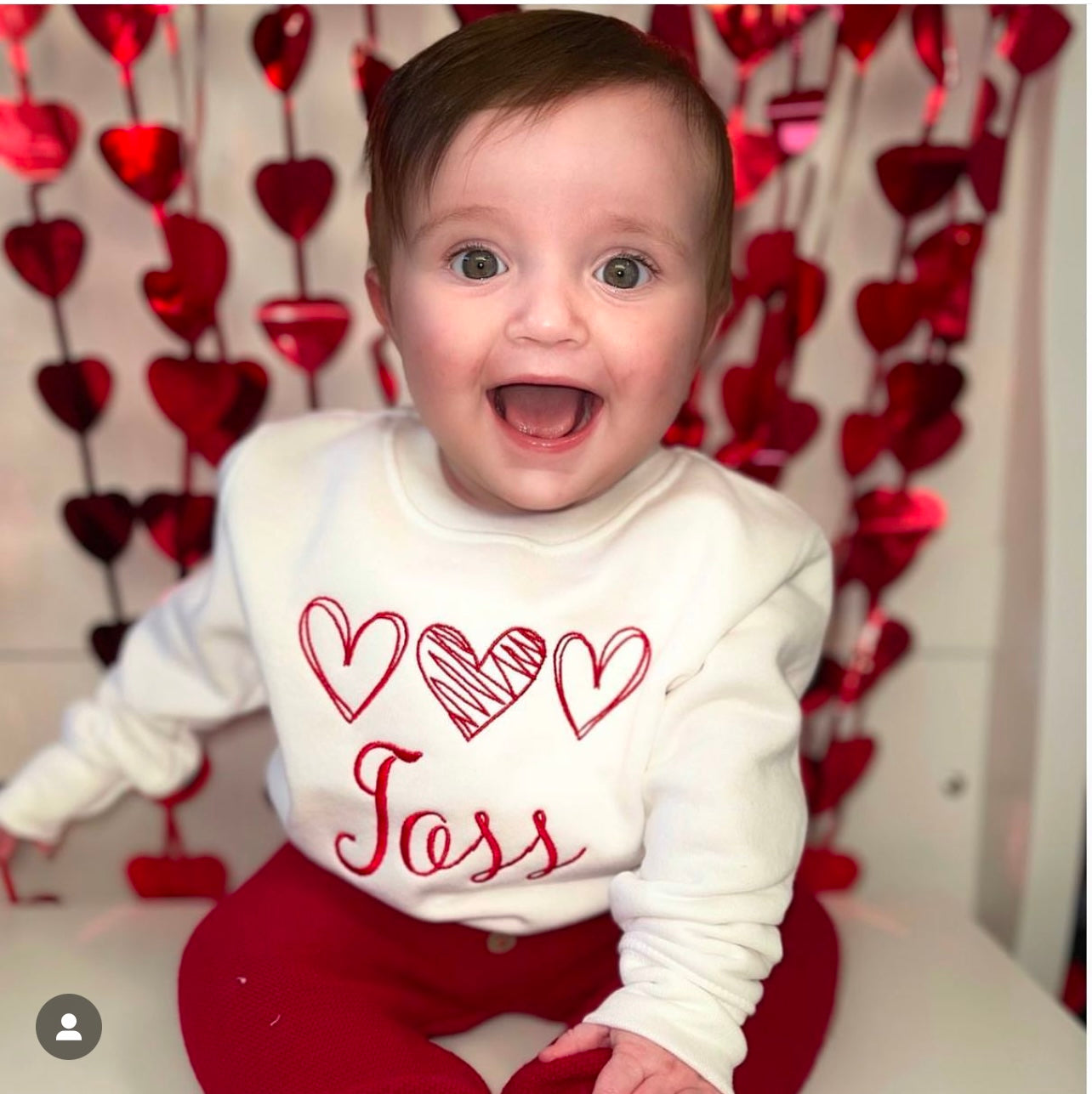 Baby wearing a white shirt with red hearts and 'Jess' text, standing in front of a heart-patterned backdrop.