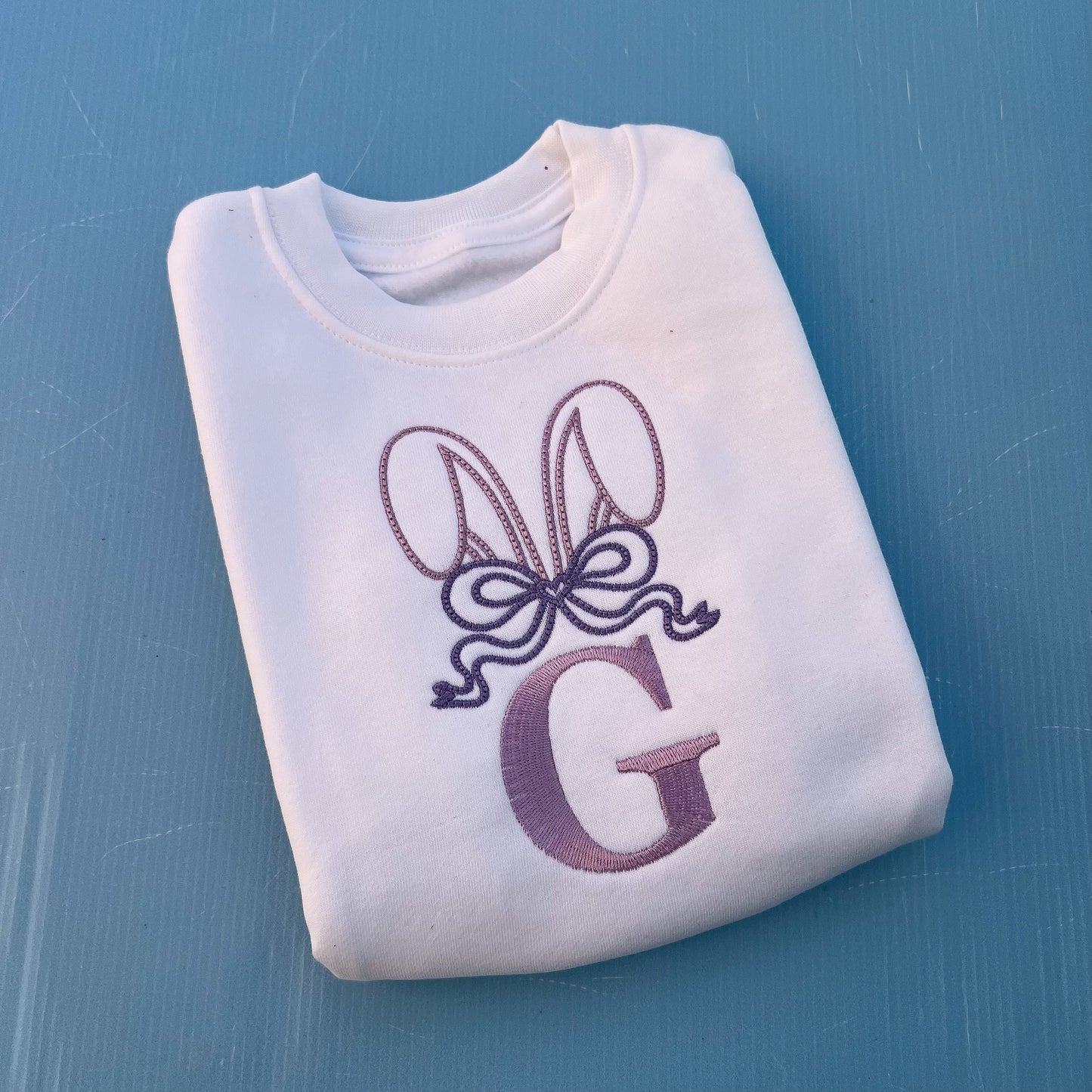 White jumper with embroidered bunny ears, a large bow and the letter G 