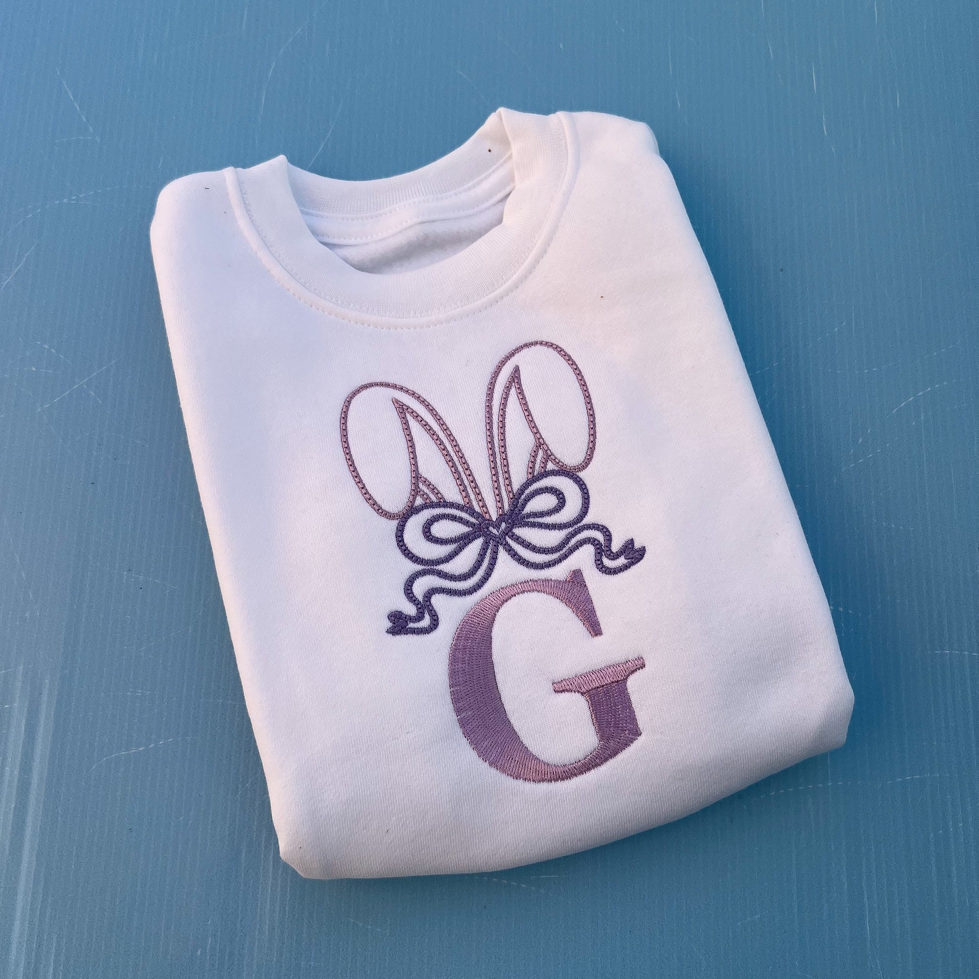 White jumper with embroidered bunny ears, a large bow and the letter G 