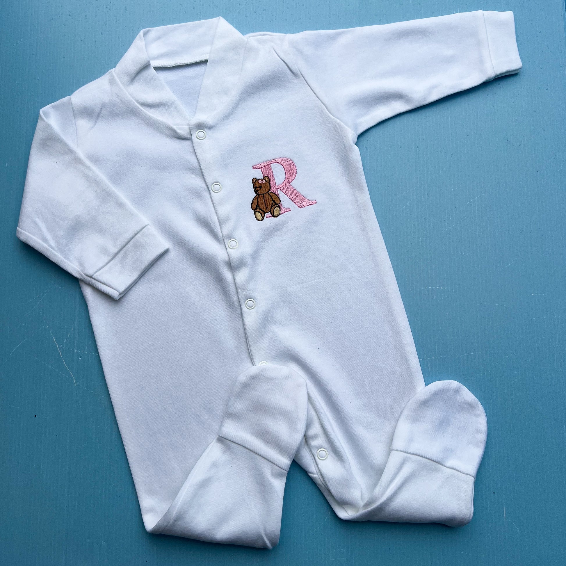 White baby onesie with a teddy bear and letter 'R' on a blue background