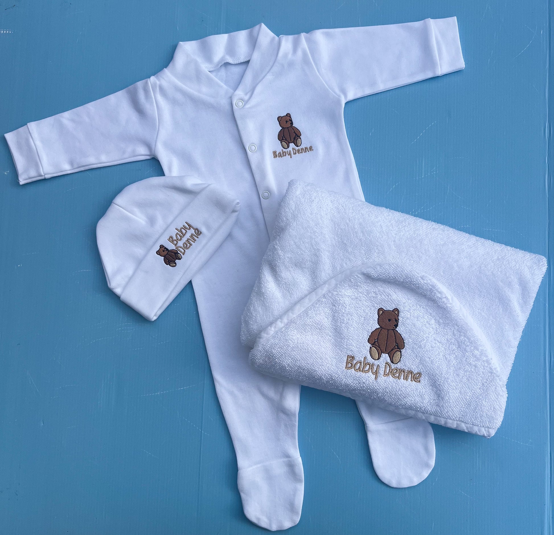 Baby clothing set with white outfit, hat, and towel featuring 'Baby Denne' branding on a blue background.