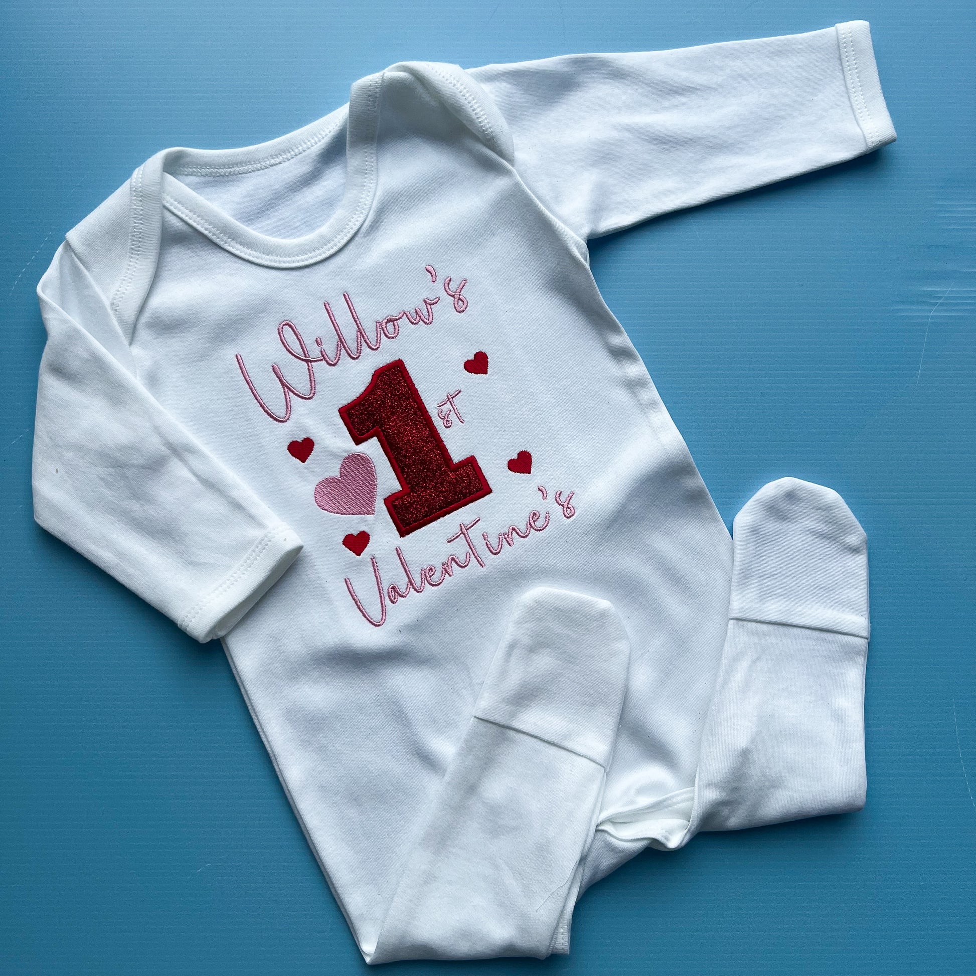 White baby onesie with 'Willow's 1st Valentine's' text on a blue background