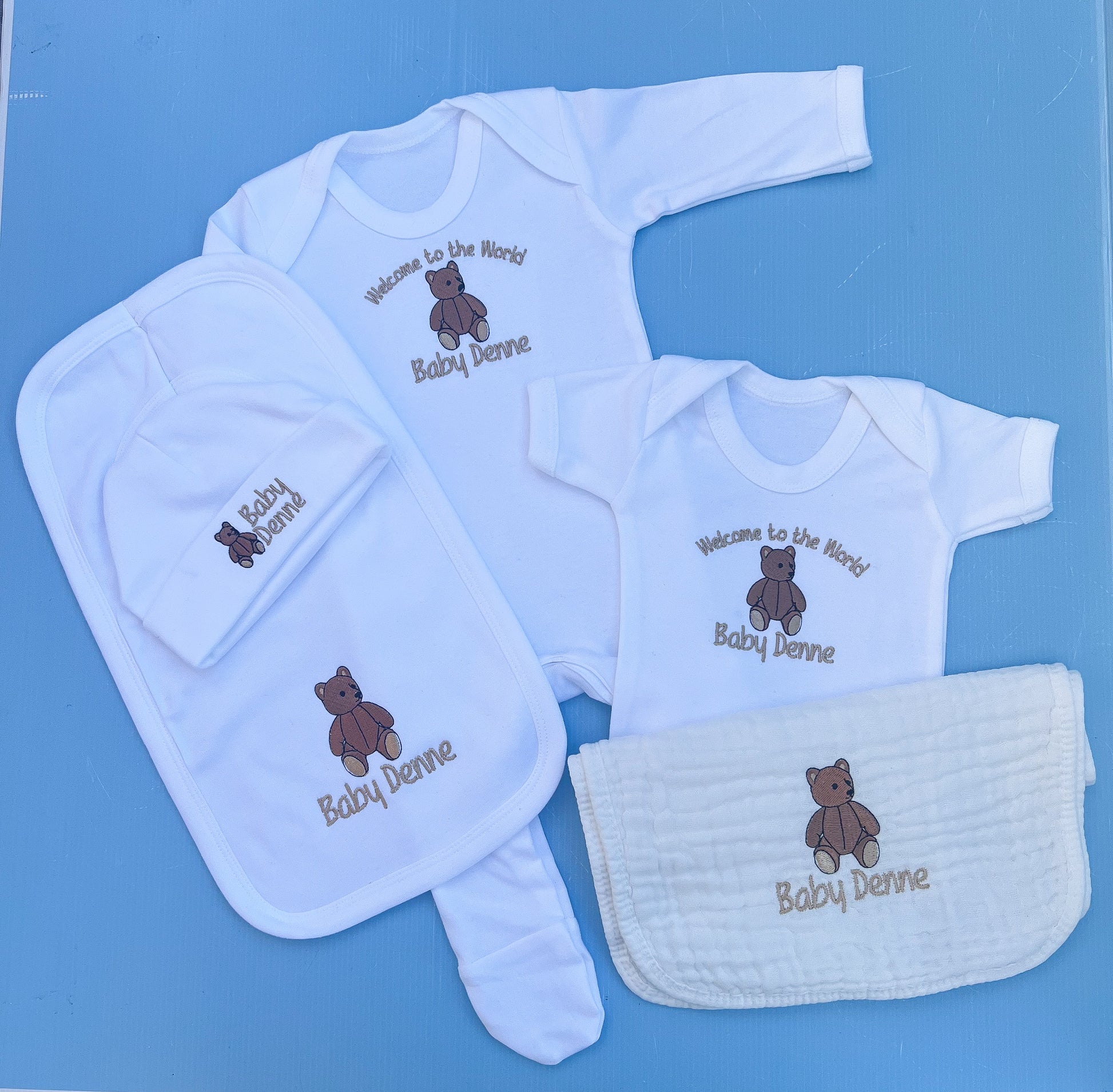 Baby clothing set with embroidered text and teddy bear design on a blue background