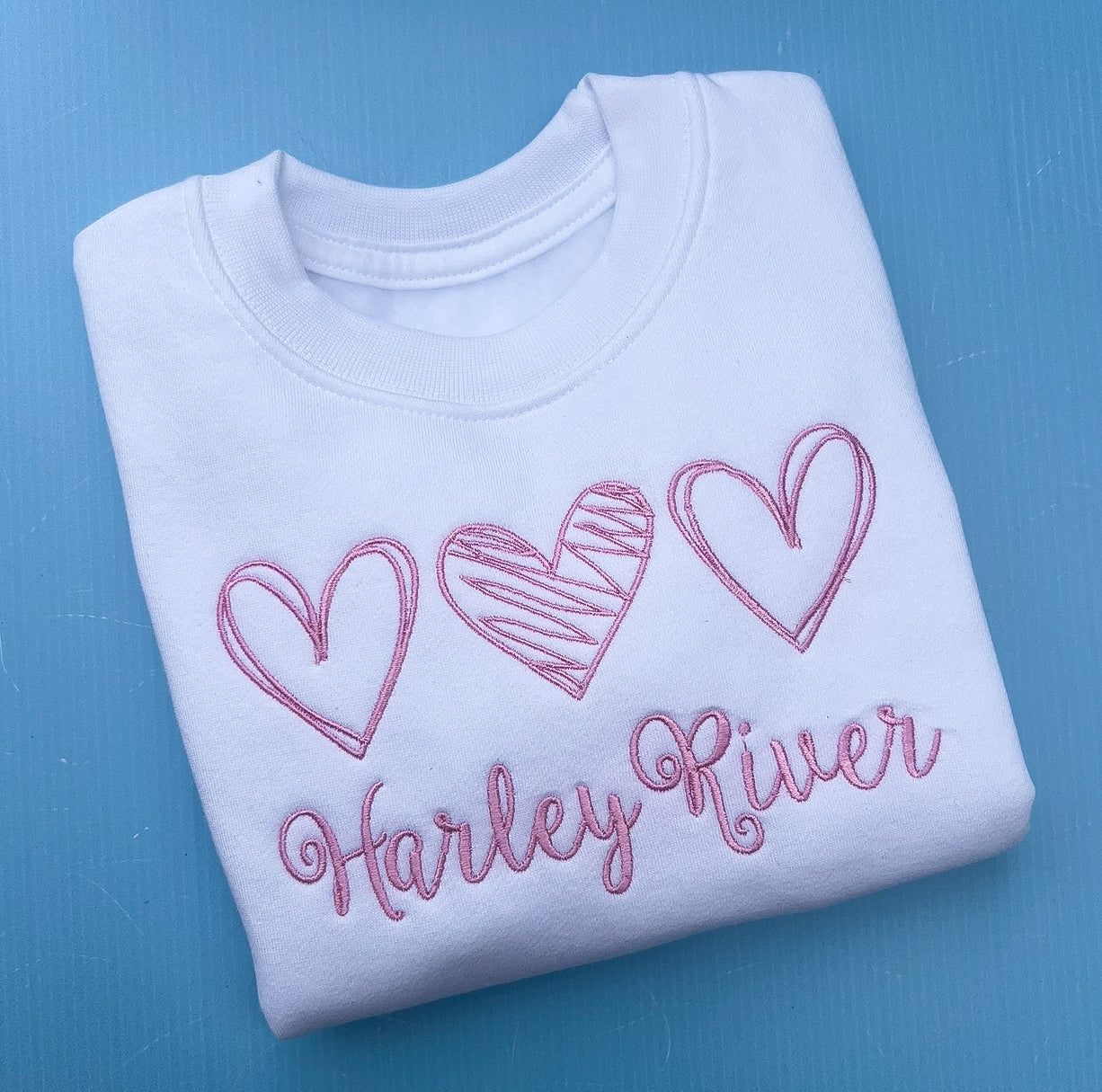 Folded white sweatshirt with pink hearts and 'Harley River' text on a blue background