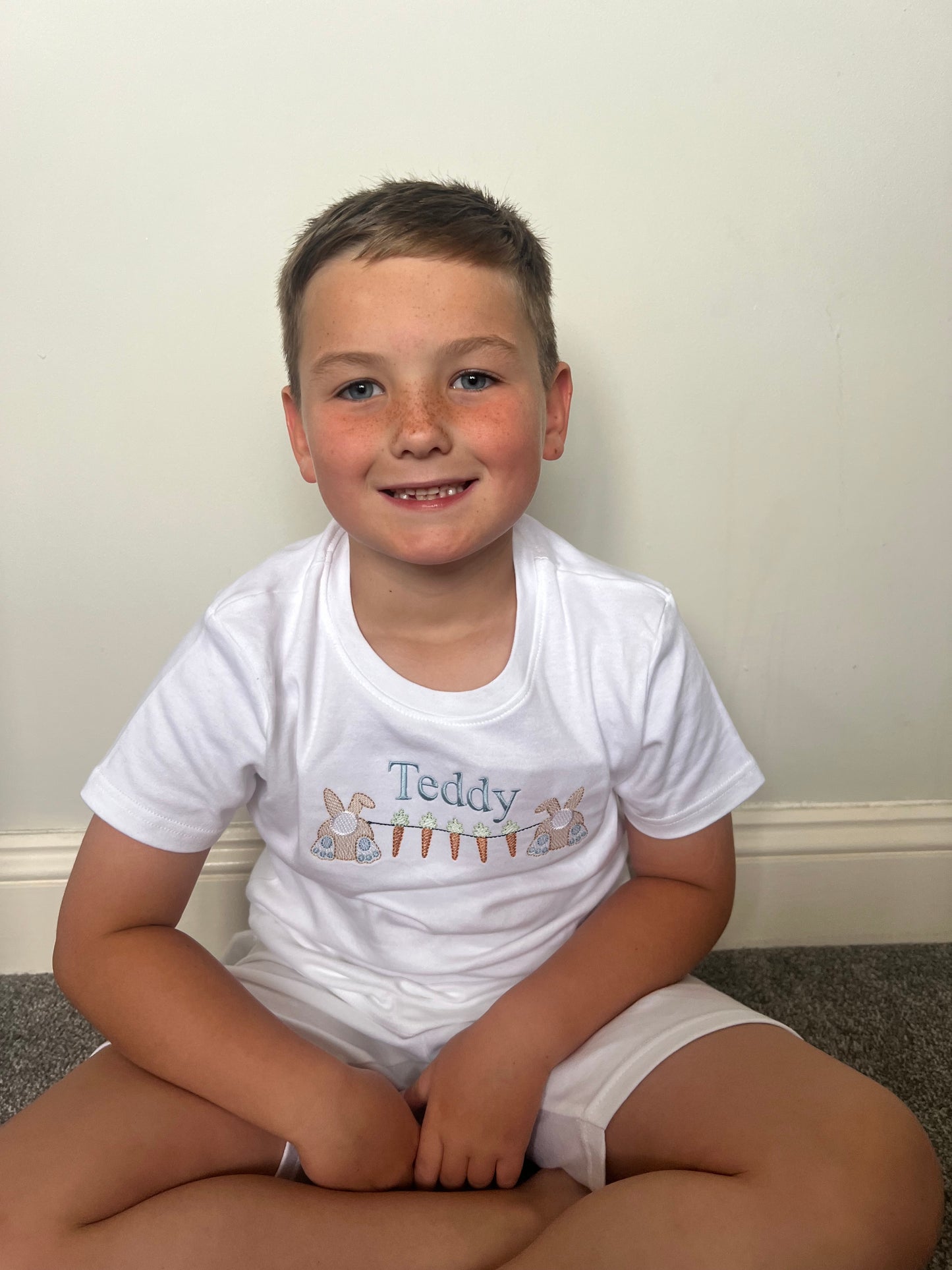Boy wearing white short pyjamas with carrot bunting embroidery on the front 