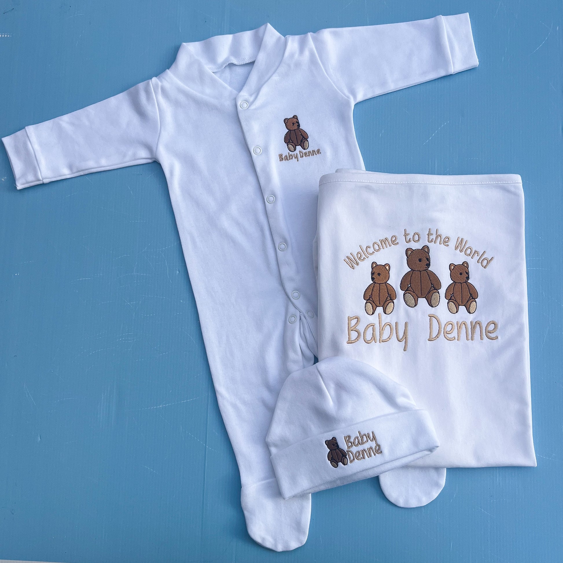 Baby outfit with white onesie, hat, and blanket featuring teddy bear design and text on a blue background.