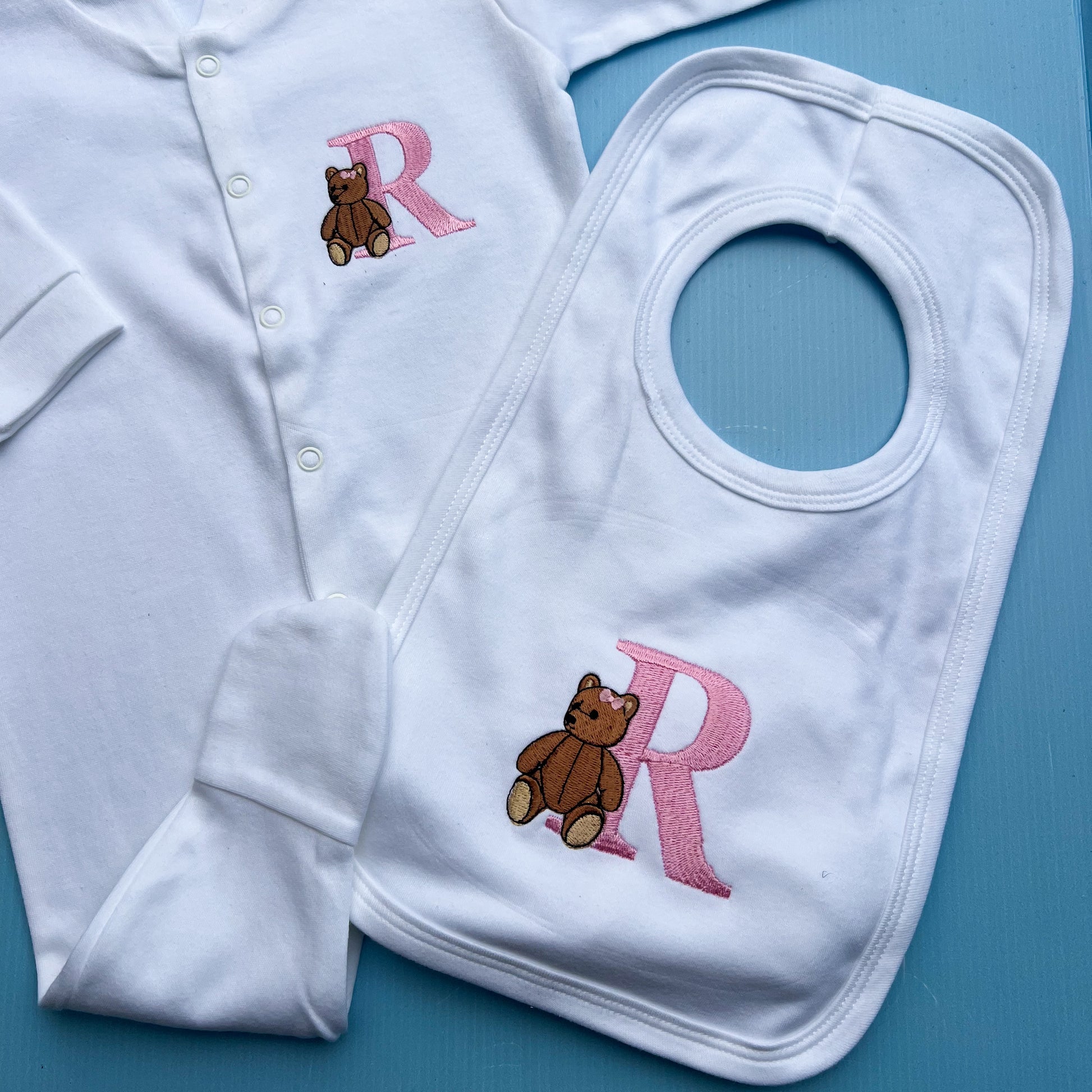 White baby outfit with embroidered bear and 'R' on a blue background