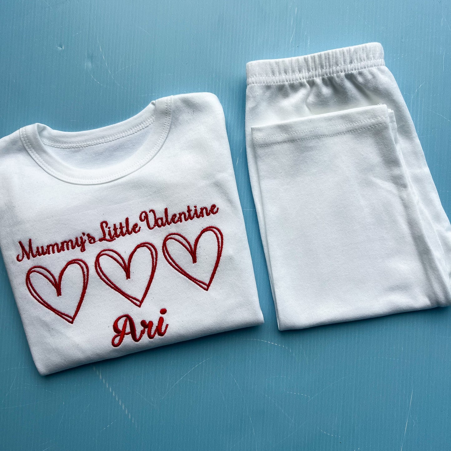 White pyjamas with 'Mummy's Little Valentine Ari' text and hearts on a blue background
