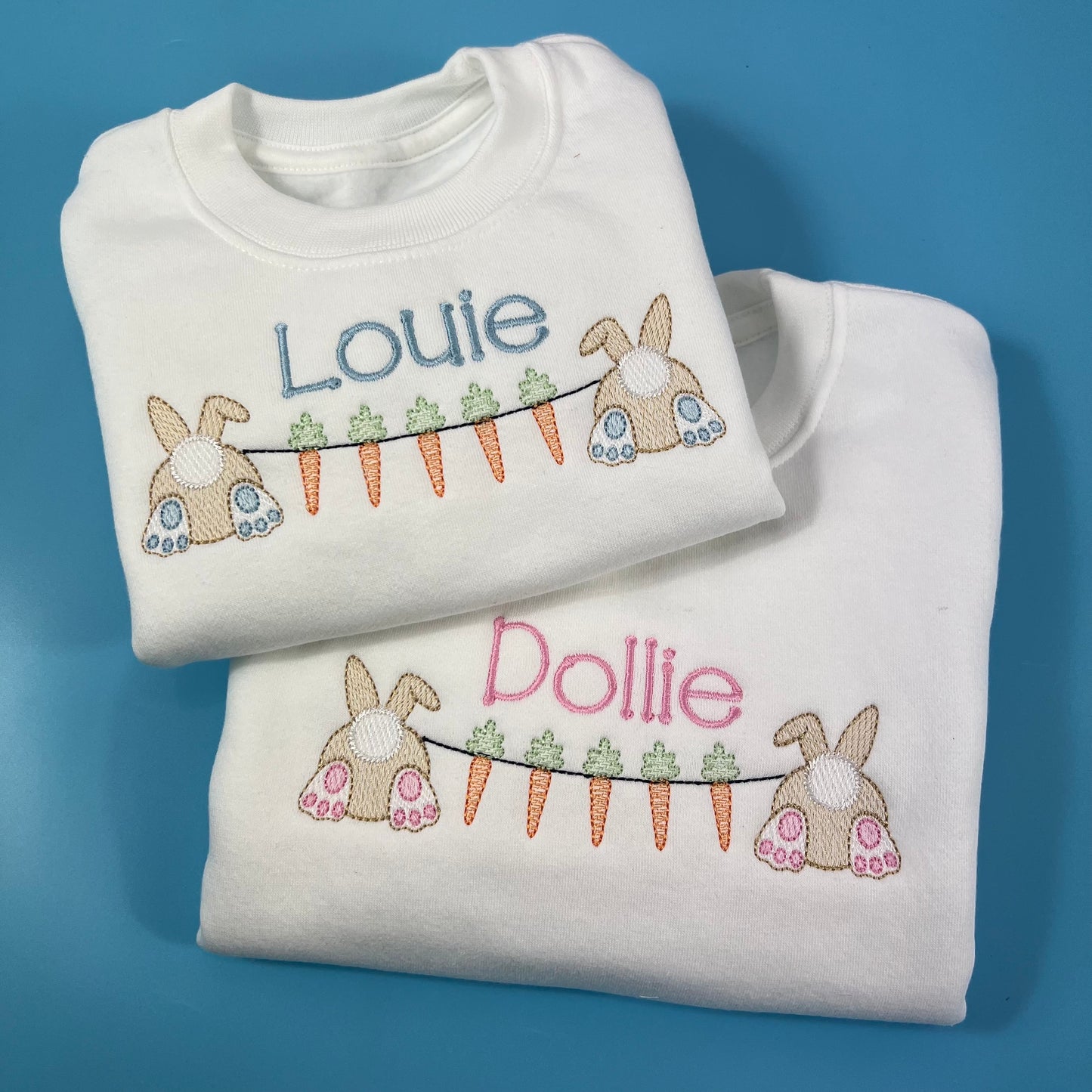 Two folded white sweatshirts with Easter bunting embroidered designs and names on a blue background