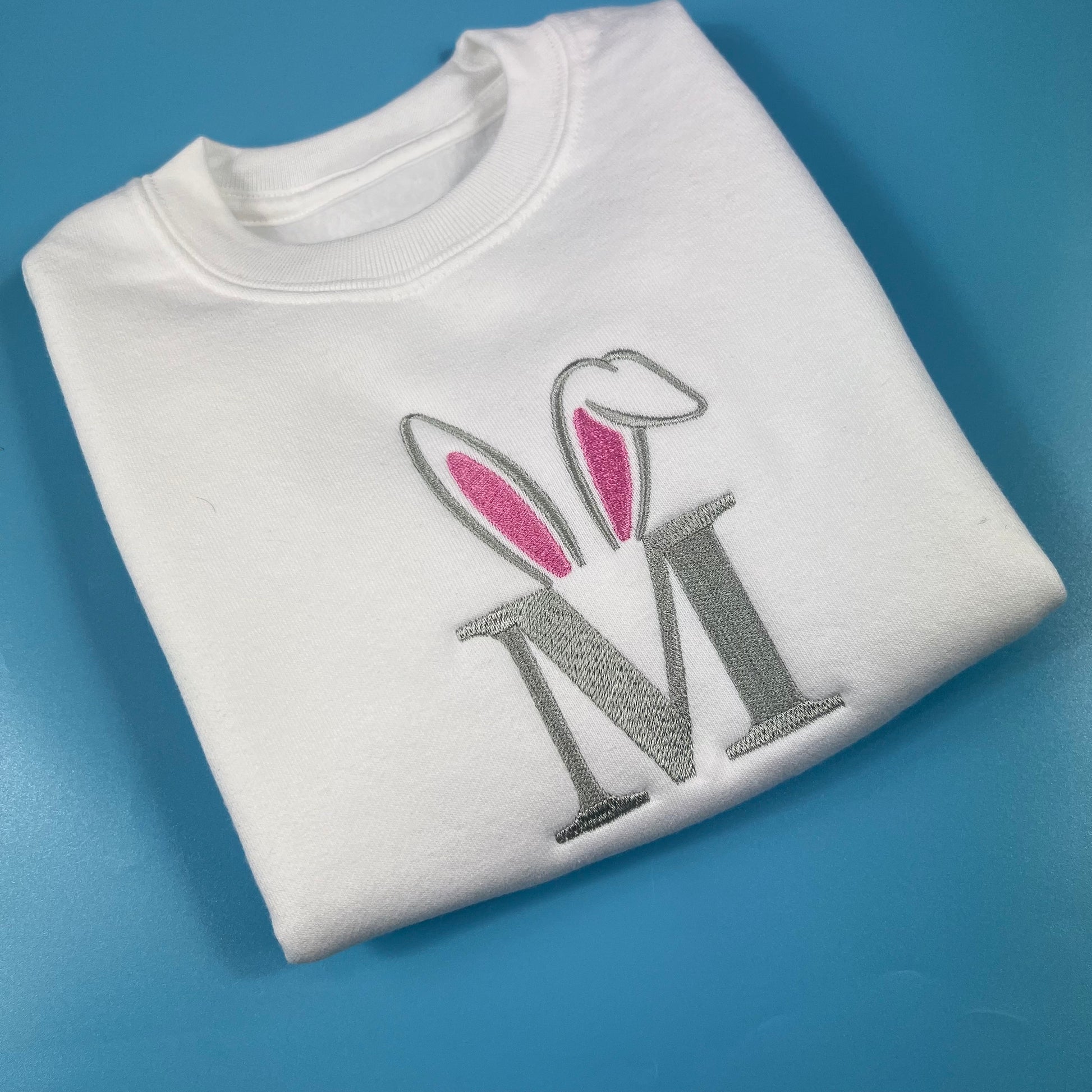 White sweatshirt with a Easter decorative letter 'M' design on a blue background