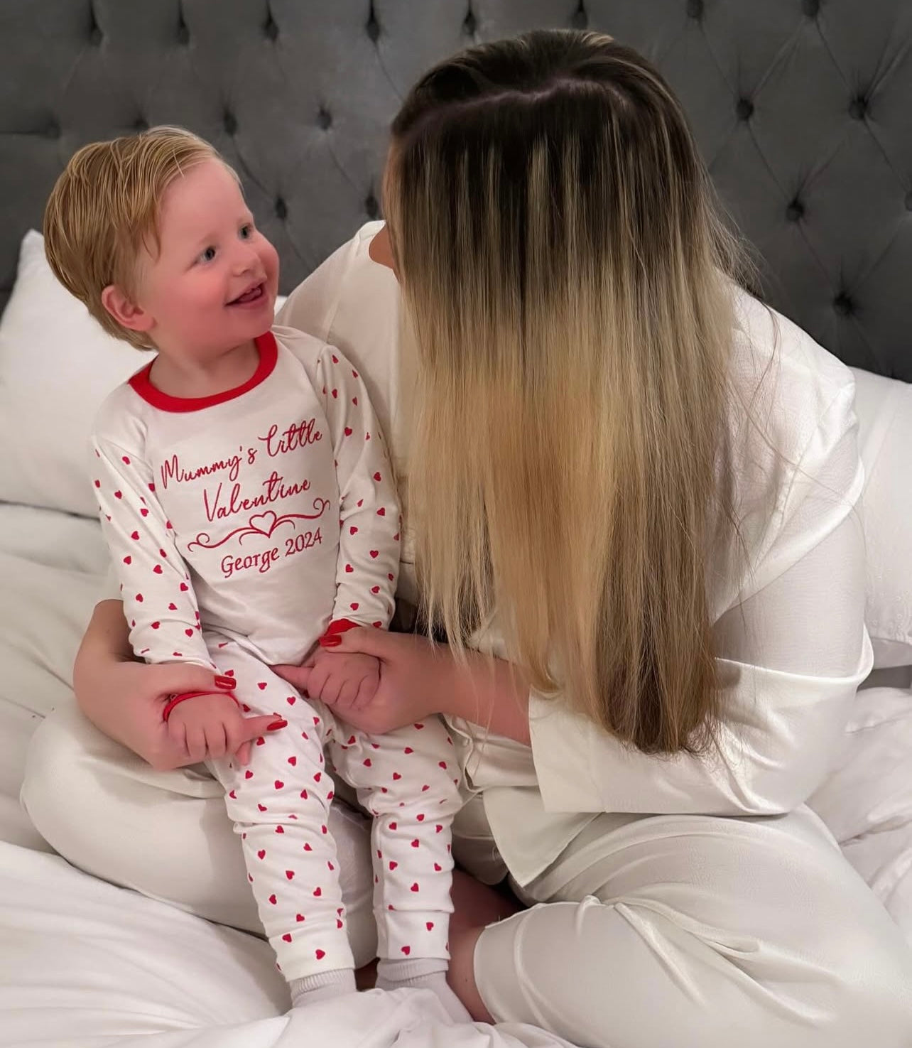 Woman and child in pajamas sitting on a bed, with the woman holding the child's hand.