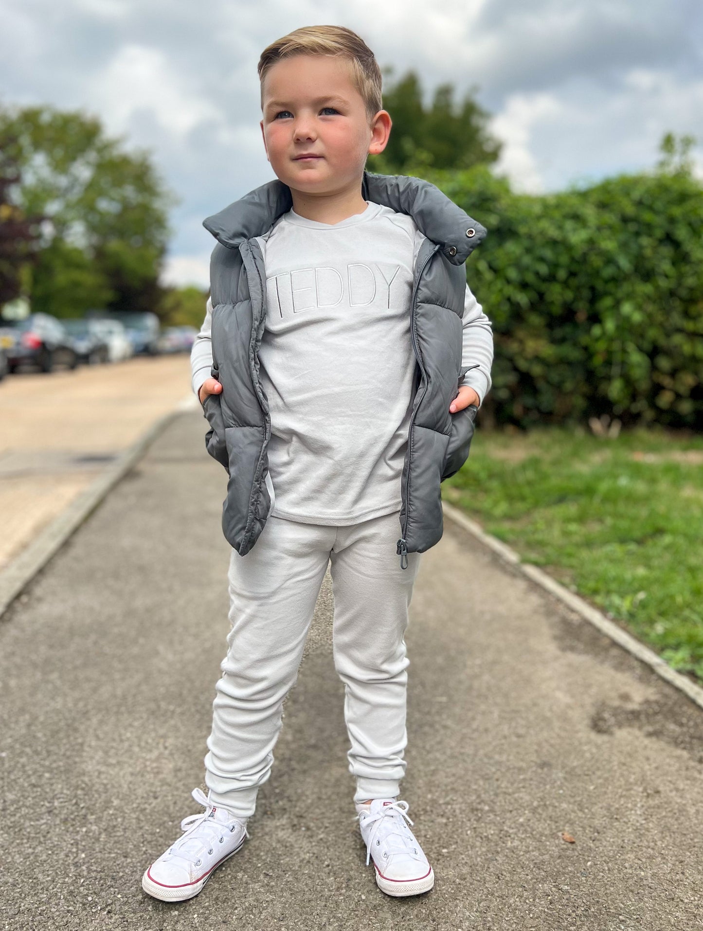 Young boy standing on a path wearing white converse and a soft grey tracksuit with cuffed ankles and sleeves, also his name embroidered in grey across the chest, child is also wearing grey body warmer