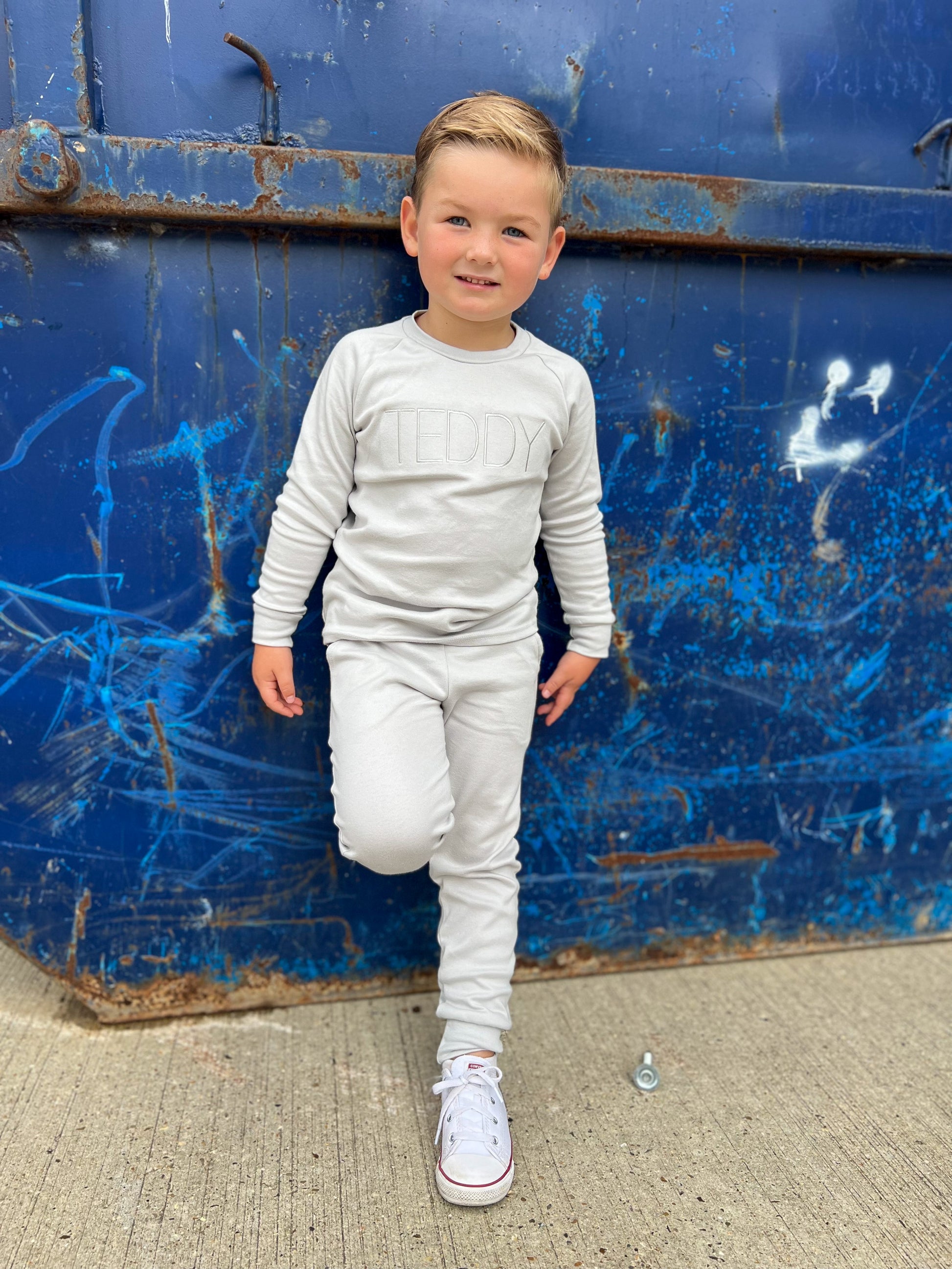 Young boy leaning against a skip wearing white converse and a soft grey tracksuit with cuffed ankles and sleeves, also his name embroidered in grey across the chest